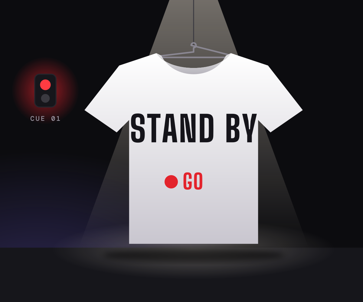 A white T-shirt printed with STAND BY and a red GO dot, hanging under a spotlight on a dark stage beside a red cue light